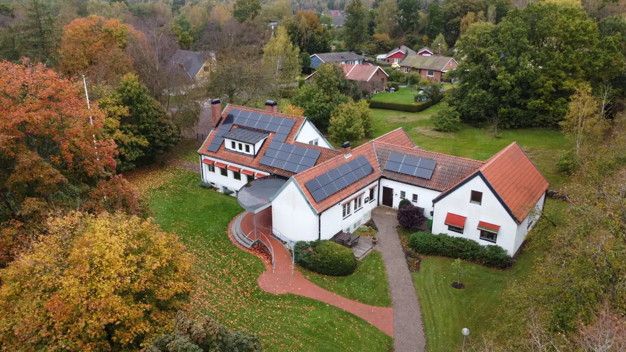 Drönarbild över vit villa med röda takpannor och solpaneler på flera takfall, omgiven av höstträd